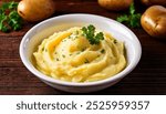 A close-up image of a white bowl filled with creamy mashed potatoes topped with fresh parsley garnish. The bowl is placed on a rustic wooden surface, with whole potatoes visible in the background. 