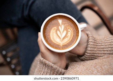 Close-up image of a woman in a cosy knitted sweater holding a cup of hot cappuccino, enjoying morning coffee while sitting at an outdoor table at a cafe in the city in the winter. - Powered by Shutterstock