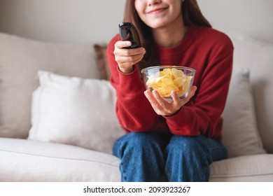 Closeup image of a woman eating potato chips while searching channel with remote control to watch tv at home