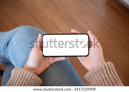 Close-up image of a woman holding a white-screen smartphone mockup in a horizontal position indoors. watching videos, playing a mobile game