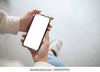 A close-up image of a woman in jeans sits indoor and uses her smartphone. A smartphone white-screen mockup in a woman's hand with a blurred background. chatting, shopping online, using mobile app - Powered by Shutterstock