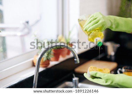 Closeup image of woman in latex gloves pouring dish soap on sponge and washing dishes