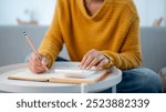 A close-up image of a woman sitting on a sofa at home, writing in a book and using a calculator to manage her household expenses and calculate her monthly savings. people and financial concepts