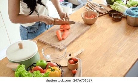 A close-up image of a woman slicing carrots on a cutting board at a wooden kitchen tabletop, cooking in the kitchen. healthy food, lifestyle, home cooking - Powered by Shutterstock