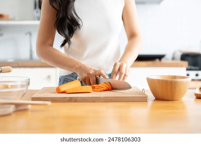 A close-up image of a woman slicing carrots on a cutting board at a wooden kitchen tabletop, cooking in the kitchen. healthy food, lifestyle, home cooking - Powered by Shutterstock