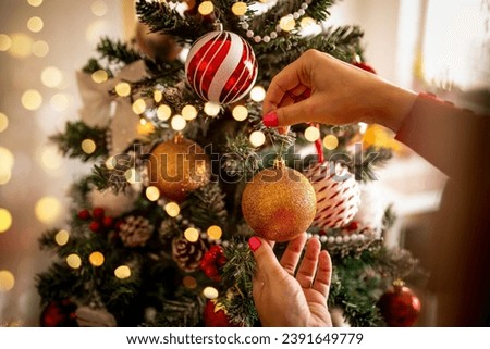 Closeup image of woman in sweater decorating Christmas tree with baubles. Woman hands holding and hanging decoration for New year