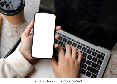 Close-up image of a woman using her smartphone while working on her laptop at her modern desk. A white-screen smartphone to display your graphic ads. People and technology concepts