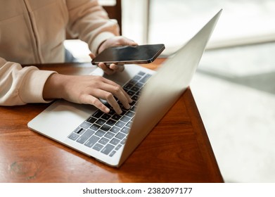 Close-up image of a woman using her smartphone while working on her laptop at a table in a cafe. typing on the keyboard, searching, or browsing the internet. People and technology concepts