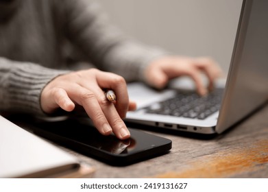 Close-up image of a woman using her smartphone while typing on a keyboard, working on her laptop computer at a table. remote job, work from home, digital nomad, wireless technology