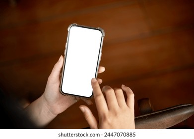 A close-up image of a woman using her smartphone indoors. A white-screen smartphone mockup in a woman's hand over a blurred rustic wood background. people and wireless technology concepts - Powered by Shutterstock