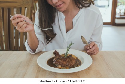 Closeup image of a woman using knife and fork to eat chicken stew on wooden table