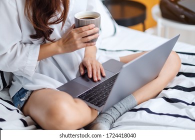 Closeup image of a woman using and working on laptop computer , drinking coffee while sitting on a white cozy bed at home