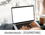 A close-up image of a woman working on her laptop computer at a table in a coffee shop, typing on the laptop keyboard. the laptop with a white screen mockup