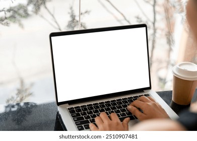 A close-up image of a woman working on her laptop computer at a table in a coffee shop, typing on the laptop keyboard. the laptop with a white screen mockup