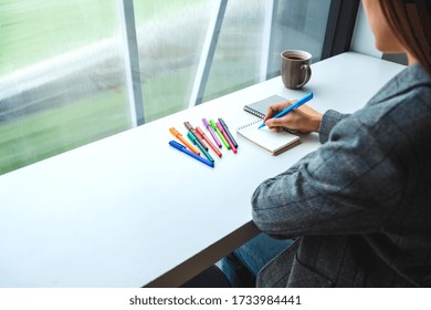 Closeup image of a woman writing on a blank notebook with colored pens and coffee cup on the table  - Powered by Shutterstock