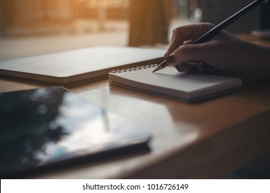 Closeup image of woman's hand writing on a blank notebook with laptop and tablet on wooden table background