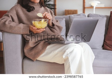 Closeup image of a young woman eating potato chips while watching on laptop computer at home