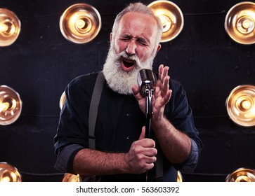 Close-up Of Impassioned Man Singing In A Silver Vintage Microphone Wearing Dark Shirt And Suspenders. Using Studio Microphone And Posing At The Camera