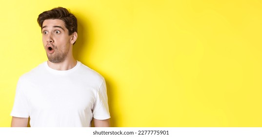 Close-up of impressed man looking left, gasping amazed, standing in white t-shirt against yellow background. - Shutterstock ID 2277775901