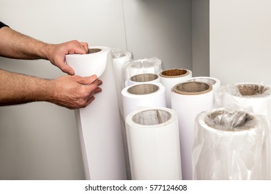 Close-up of incognito male hands choosing roll of white paper for wide format printer, from bunch of rolls in corner of the room