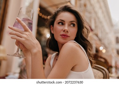 Close-up of incredibly image of pretty caucasian young girl with glass of delicious drink. Brunette with wavy hair sitting in street cafe looks to side dressed in white top.