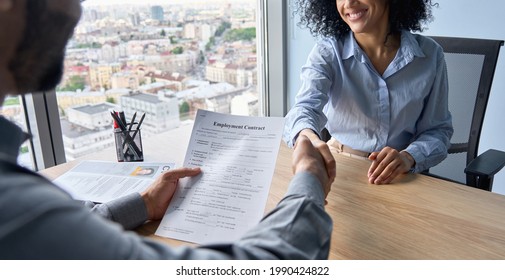 Closeup of Indian employer hr holding employment contract job offer hiring welcoming female African newcomer worker manager shaking hands in contemporary office. Getting new job handshake concept.