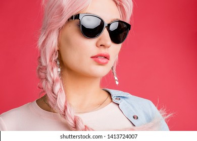 Closeup Indoor Shot Of Caucasian Woman With Pink Braid Wearing Sunglasses, Earrings Pale Pink T Shirt Looking At Camera With Open Mouth On Coral Background. Summer, Beauty, Hair Dye, Cosmetics Concept