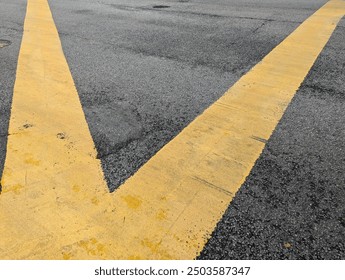 A close-up of intersecting yellow road lines on an asphalt surface, forming a V-shape. The worn texture of the paint and roughness of the asphalt highlight the urban environment and road maintenance.