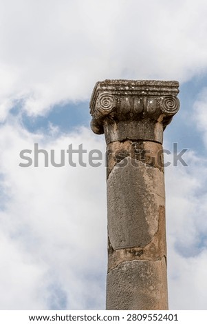 A close-up of an Ionic column capital at Volubilis shows the characteristic scroll volutes and acanthus detailing, weathered by centuries, against a bright cloudy sky.