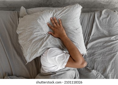 Closeup of irritated young black man lying in bed and covering head and ears with pillow, hearing and suffering from too loud sound. Angry millennial guy can't sleep, tired of noisy neighbors
