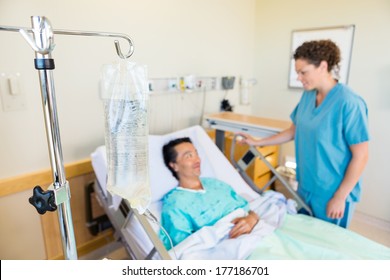 Closeup Of IV Bag With Nurse And Patient Looking At Each Other In Background At Hospital