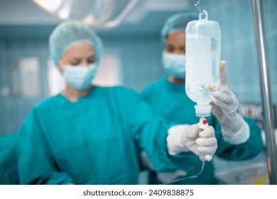 Closeup of IV drip in operating room. Female surgeon and nurse preparing intravenous drip for patient in operating theatre. Focus is on the IV drip. - Powered by Shutterstock