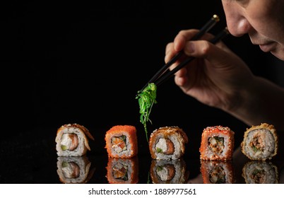 Close-up Japanese chef with seaweed on chopsticks serves assorted colourful rolls stand in row on black glossy dish. - Powered by Shutterstock