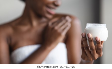 Closeup of jar with body lotion or cream in unrecognizable naked african american lady hand, black woman applying skin care product on her shoulder after shower, panorama. Soft skin concept