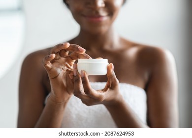 Closeup of jar with body lotion or cream in unrecognizable naked african american lady hand, black woman applying skin care product on her skin after shower, cropped. Soft skin concept