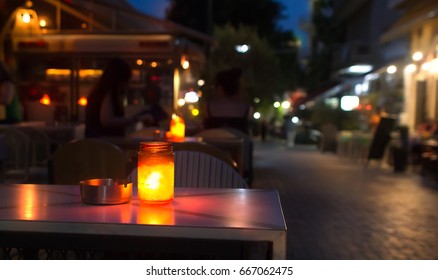 Closeup Of A Jar With A Candle On A Table With An Ashtray, Placed Outside A Bar At A Pedestrian Street In Greece. Summer Evening.