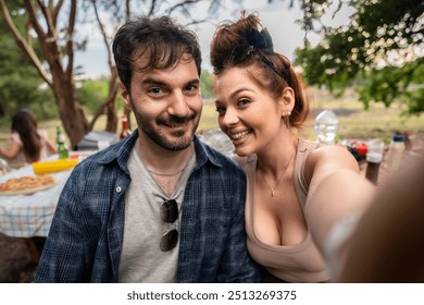 Close-up of a joyful couple taking a selfie at an outdoor picnic. Young man and woman smiling at camera, with picnic table and nature in background, capturing a moment of happiness and togetherness. - Powered by Shutterstock