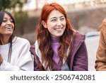 Close-up of a joyful young woman with red hair, smiling broadly while spending time with her diverse group of friends. Authentic portrait of genuine happiness and friendship in an outdoor setting.