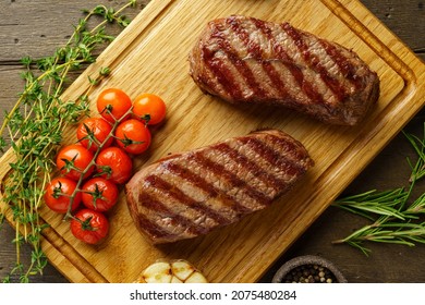 Closeup Of Juicy And Tender Top Blade Beefsteaks On Wooden Board With Rosemary, Thyme, Roasted Garlic Clove, Pepper, Cherry Tomatoes And Red Chili. Grilled Meat With Herbs And Spices. Gourmet Keto