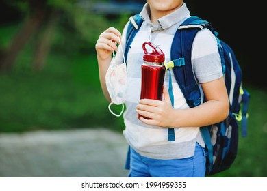 Closeup Of Kid Boy, Medical Mask, Water Bottle And Backpack Or Satchel. Schoolkid On Way To School. Child Outdoors. Back To School After Quarantine Time From Corona Pandemic Disease Lockdown. No Face.