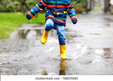 Close-up Of Kid Wearing Yellow Rain Boots And Walking During Sleet, Rain And Snow On Cold Day. Child In Colorful Fashion Casual Clothes Jumping In A Puddle. Having Fun Outdoors.