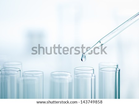 A close-up of a laboratory glass pipette with emerging drop of substance over one of several test tubes on a light  background