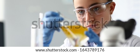 Close-up of laboratory researcher holding medical glass bottle with yellow liquid wearing sterile gloves and special lab eyewear. Scientist woman at job. Chemistry concept