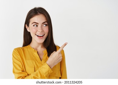 Close-up of lady smiling, pointing and looking right with surprised face, standing on white background