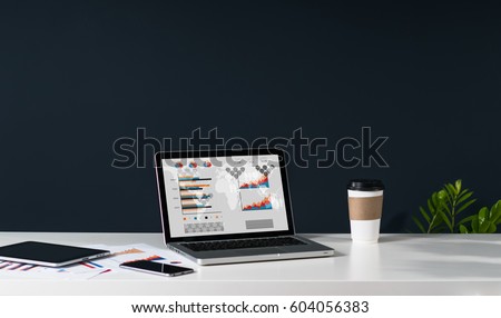 Close-up of laptop with graphs, charts and diagrams on screen on white table. Nearby is tablet computer, smartphone, paper graphics, cup of coffee. In background dark wall. Empty workplace.