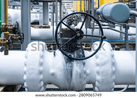 Close-up of a large manual gate valve on frozen pipelines covered in ice due to cryogenic gas flow inside an LNG or industrial plant surrounded by yellow-painted steel structures.