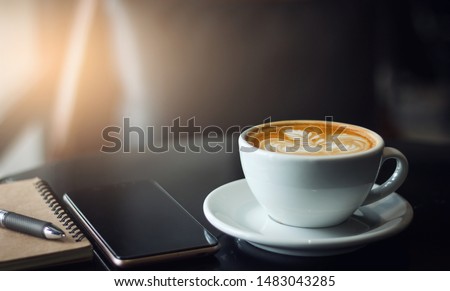 Closeup latte art coffee in white cup with smartphone and notebook on black table. Vintage color tone