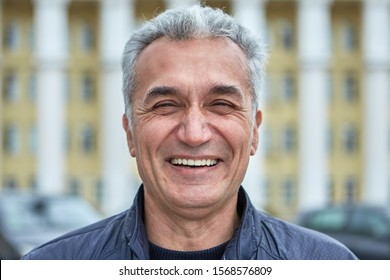 Closeup Laughing Face Of An Elderly Businessman, Civil Servant, Doctor Or Television Presenter. Portrait Of A Joyful Caucasian Man Over Fifty Years, With Short Gray Hair Outdoors, Near Public Building