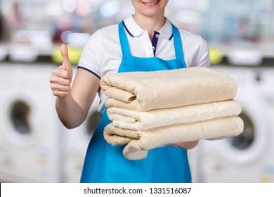 A closeup of a laundry worker holding clean towels and showing a positive gesture at the blurred laundry background. The concept of cleaning service popularity.