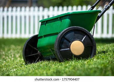 Closeup Of Lawn Spreader In Grass Yard Used For Applying Grass Seed, Fertilizer, Herbicides, Pesticides To Kill Weeds, White Picket Fence In Background, Lawn Maintenance Program, Healthy Lawn Care